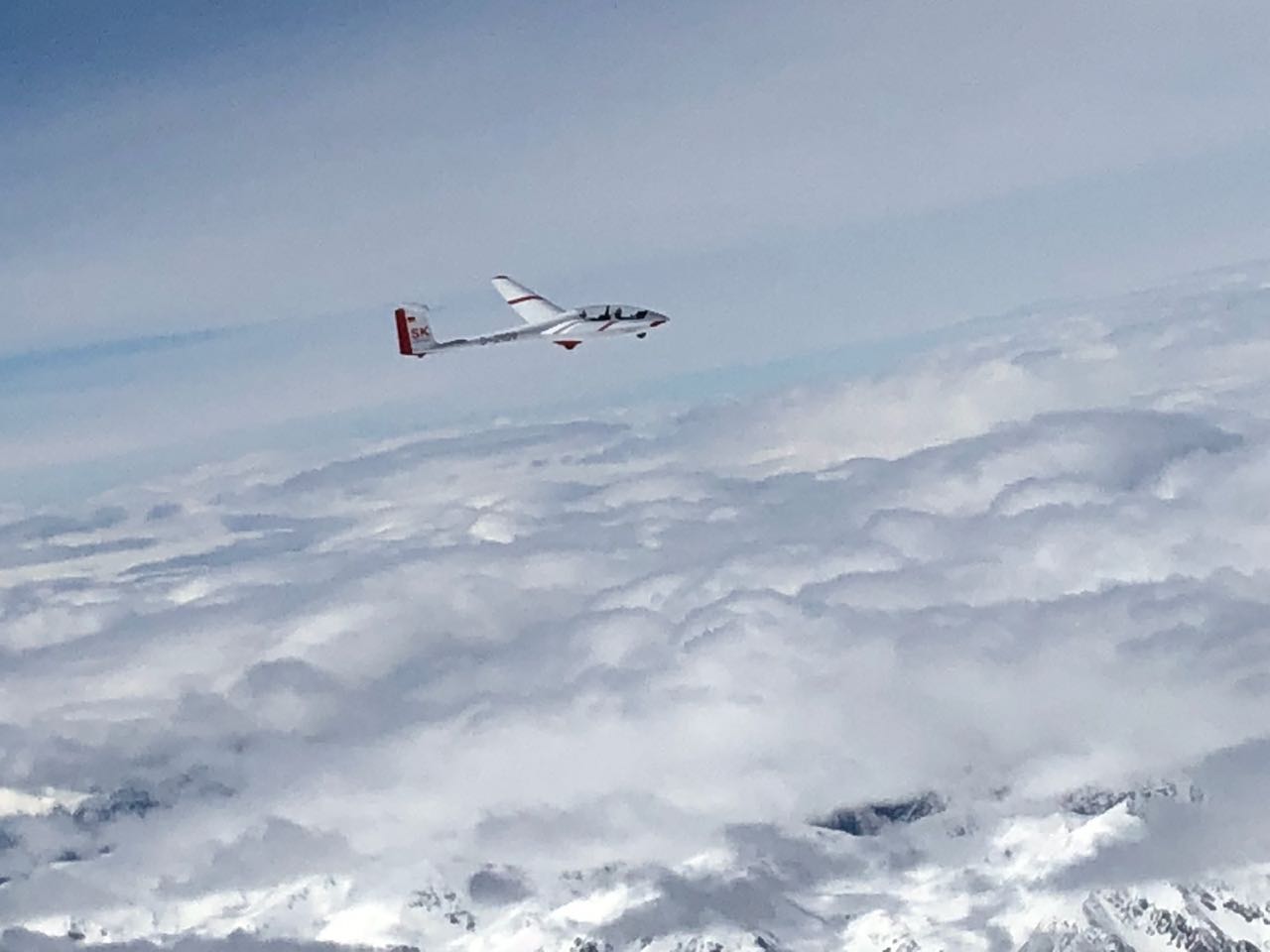 Unser jüngstes Segelflugzeug, die ASK 21 Baujahr 2017 über den Wolken bei einer Exkursion ins Hochgebirge. Eine ASK 21 mit roten Warnmarkierungen oberhalb der fast geschlossenen Wolkendecke. Im Vorderungd sieht man schneebedeckte Berggipfel durch die Wolkenlücken.