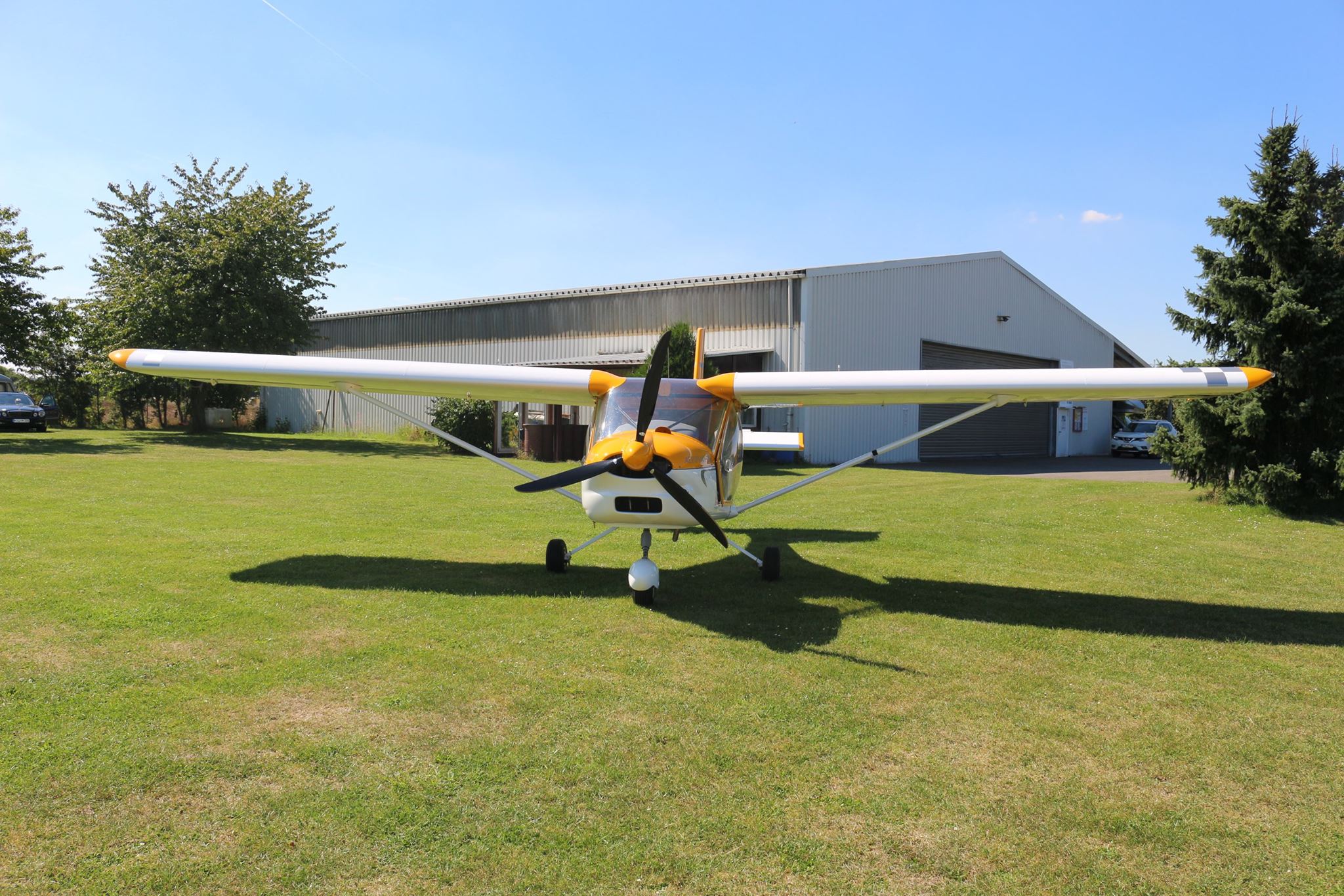 Utraleichtflugzeug AL22 vor der neuen Halle Gelb-weißes Ultraleichtflugzeug in Hochdeckerbauform auf der Wiese vor einem weißen Flugzeughangar.